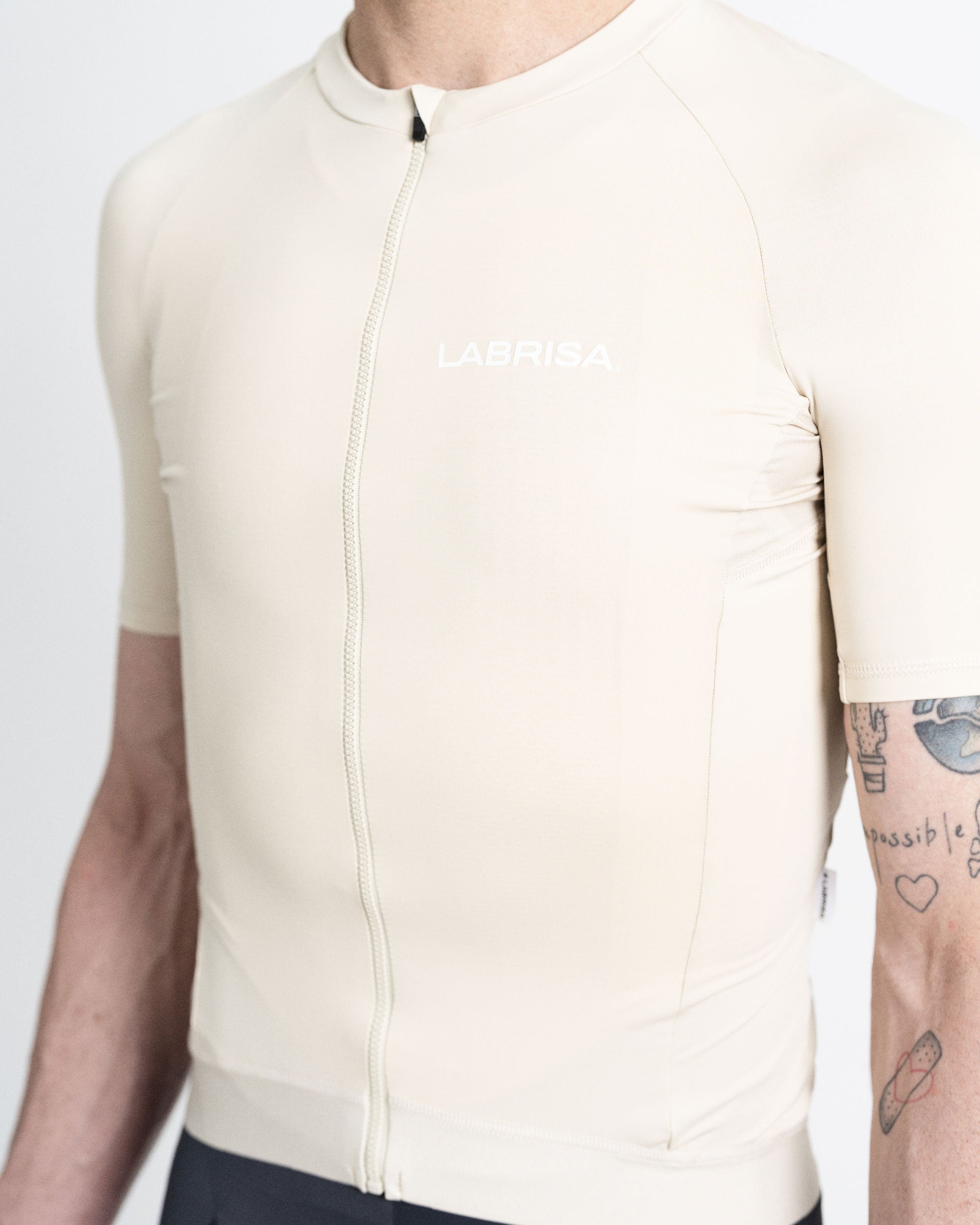 Imagen de maillot de ciclismo de mujer crema en modelo visto de frente. Logotipo de LABRISA en blanco en el pecho izquierdo e isotipo en blanco de la B en el bíceps derecho. Cremallera, siliconas y demás detalles al mismo tono crema.