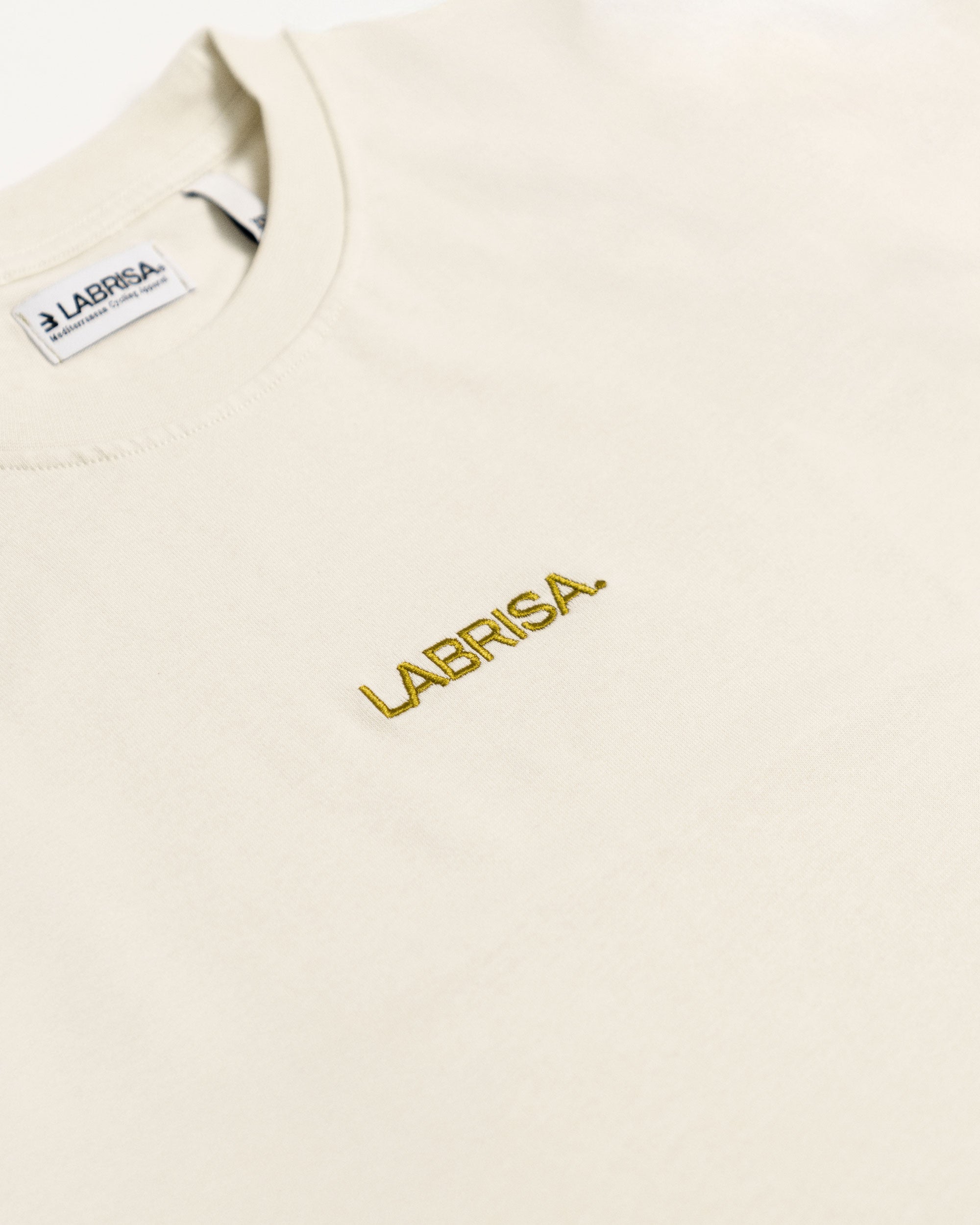 Imagen de detalle de la parte delantera de la camiseta de manga corta crema. Se ve el logotipo bordado en oliva en pecho en la parte central de la camiseta.