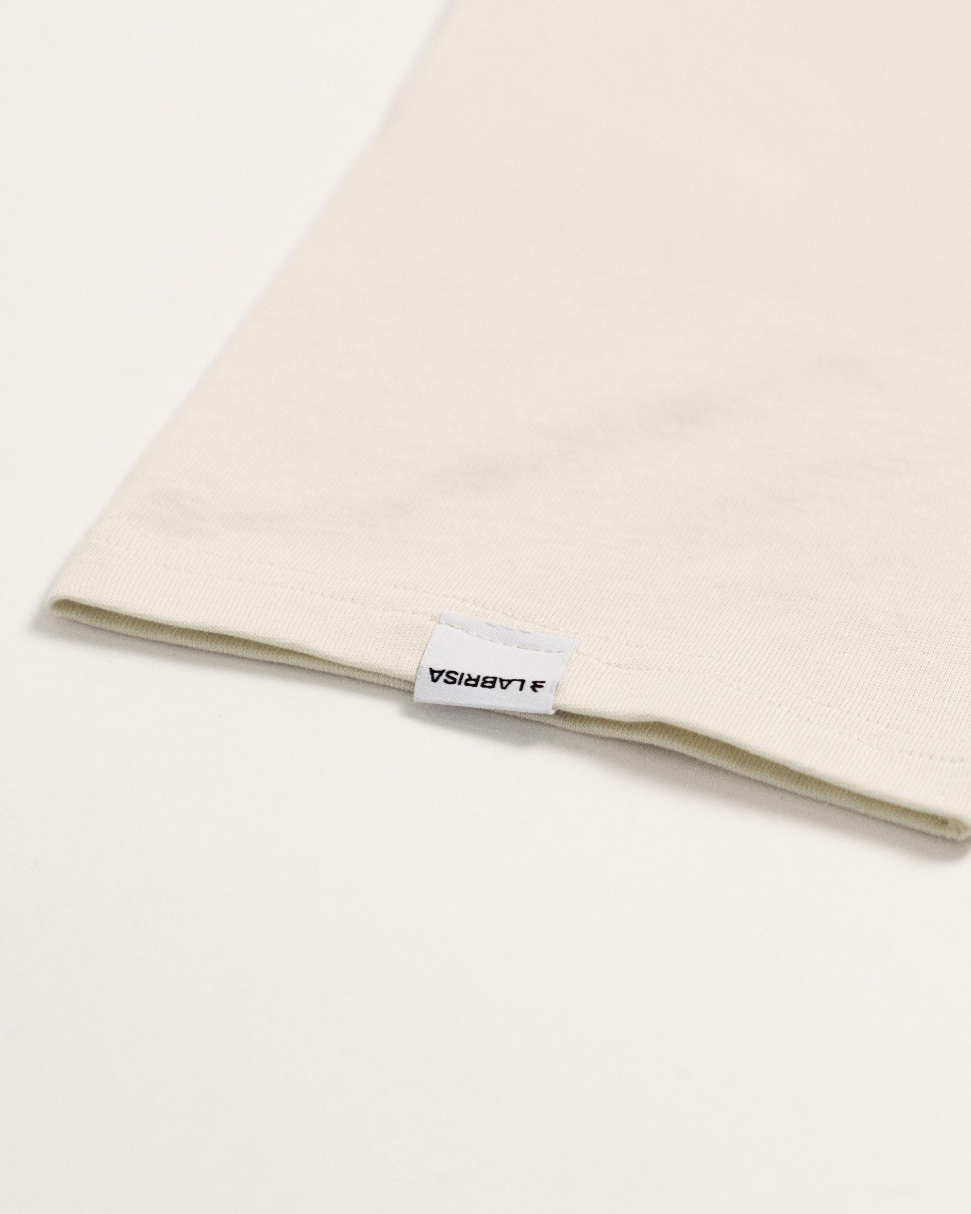Imagen de detalle de la manga de la camiseta de manga corta crema. Se ve la etiqueta tejida en la que se lee en negro sobre blanco LABRISA y está cosida en el centro de la terminación de la manga.