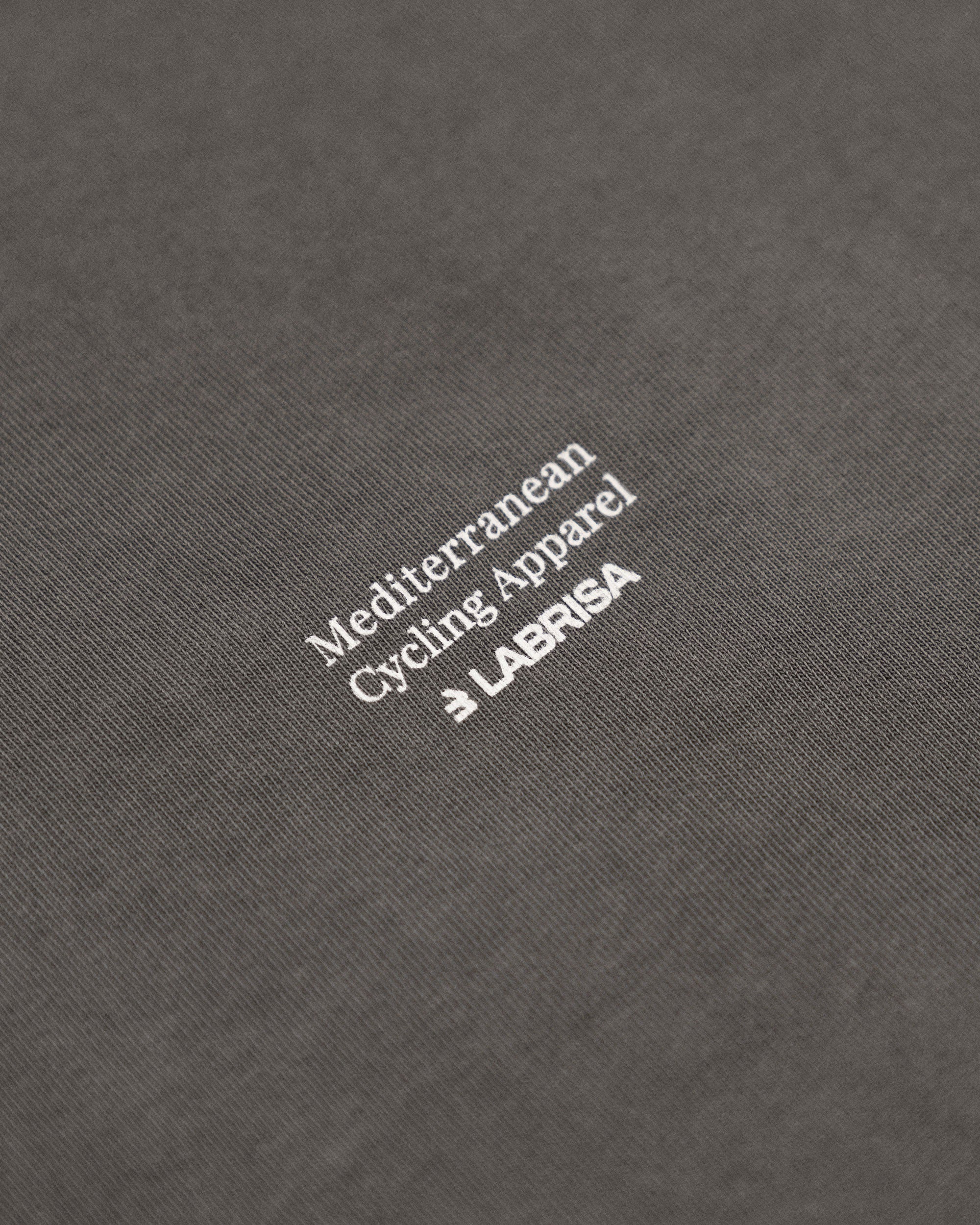 Imagen de detalle de la parte trasera de la camiseta de manga corta gris. Se ve el tagline Mediterranean Cycling Apparel en crema que va colocado en pequeño en el centro de la parte de atrás de la camiseta.