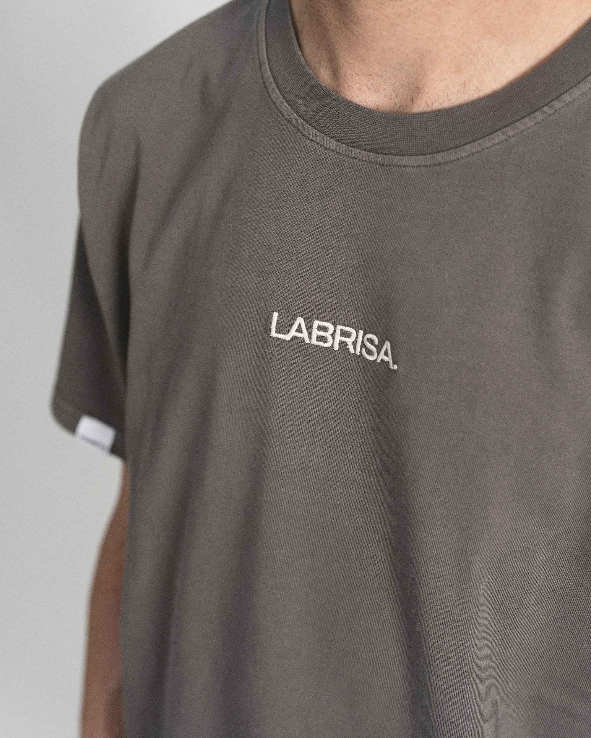 Imagen de detalle de la parte delantera de la camiseta de manga corta gris. Se ve el logotipo bordado en crema en pecho en la parte central de la camiseta.
