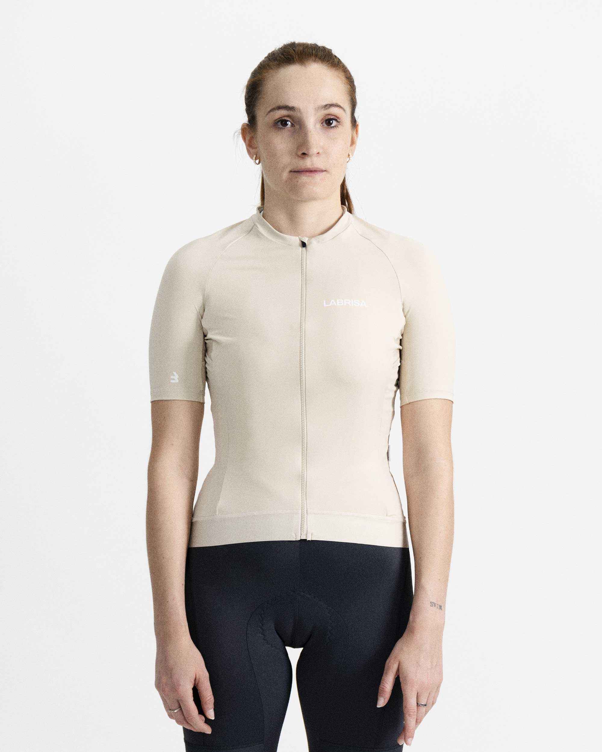 Imagen de maillot de ciclismo de mujer crema en modelo visto de frente. Logotipo de LABRISA en blanco en el pecho izquierdo e isotipo en blanco de la B en el bíceps derecho. Cremallera, siliconas y demás detalles al mismo tono crema.
